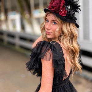 May include: A black witch hat adorned with deep red fabric flowers, black feathers, and a delicate netting veil. The hat features a pointed crown and a wide brim, accented with dark decorative elements that provide a textured appearance.