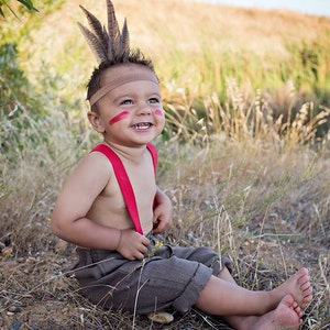 May include: A young child wearing a brown and beige outfit with red suspenders and a feather headdress. The child has red paint on their face and is smiling.