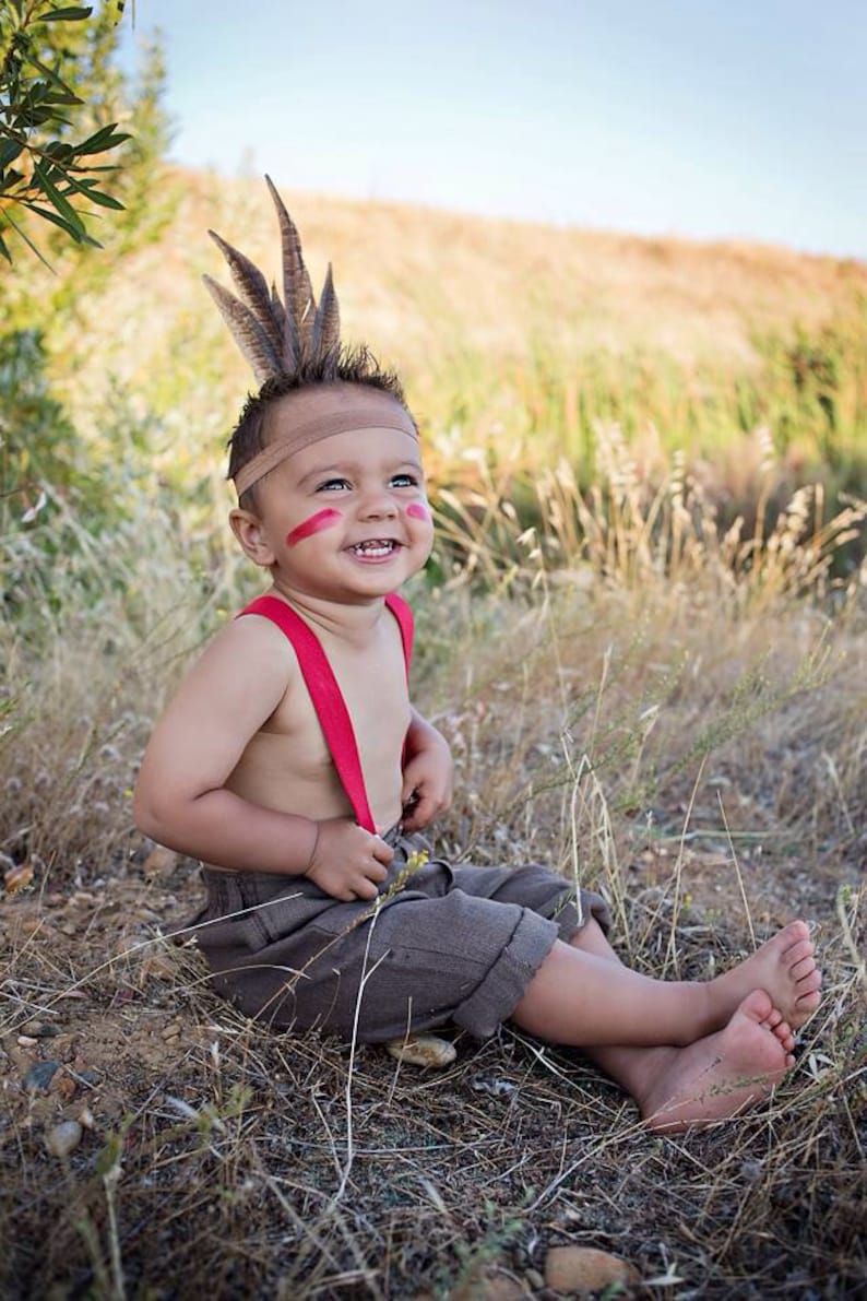 May include: A young child wearing a brown and beige outfit with red suspenders and a feather headdress. The child has red paint on their face and is smiling.