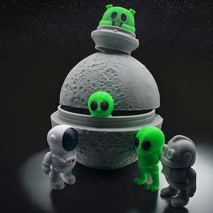 Peut inclure: Un récipient gris en forme de lune texturée avec une figurine d'extraterrestre verte dessus et à l'intérieur. Trois petites figurines d'un astronaute, d'un extraterrestre vert et d'un robot gris se tiennent devant la lune. Le fond est une scène spatiale sombre.