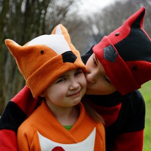 May include: Two children wearing animal-themed hats. The child on the left is wearing an orange fox hat and a matching orange jacket. The child on the right is wearing a red and black raccoon hat and a red and black jacket. The child on the right is kissing the child on the left on the cheek.
