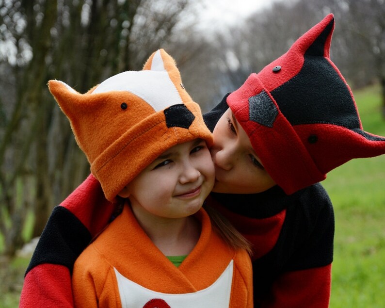 May include: Two children wearing animal-themed hats. The child on the left is wearing an orange fox hat and a matching orange jacket. The child on the right is wearing a red and black raccoon hat and a red and black jacket. The child on the right is kissing the child on the left on the cheek.