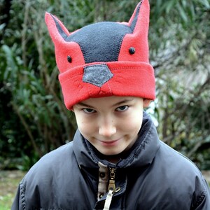 May include: A child wearing a red and black fox hat with a mischievous expression. The hat has large ears and a black nose.