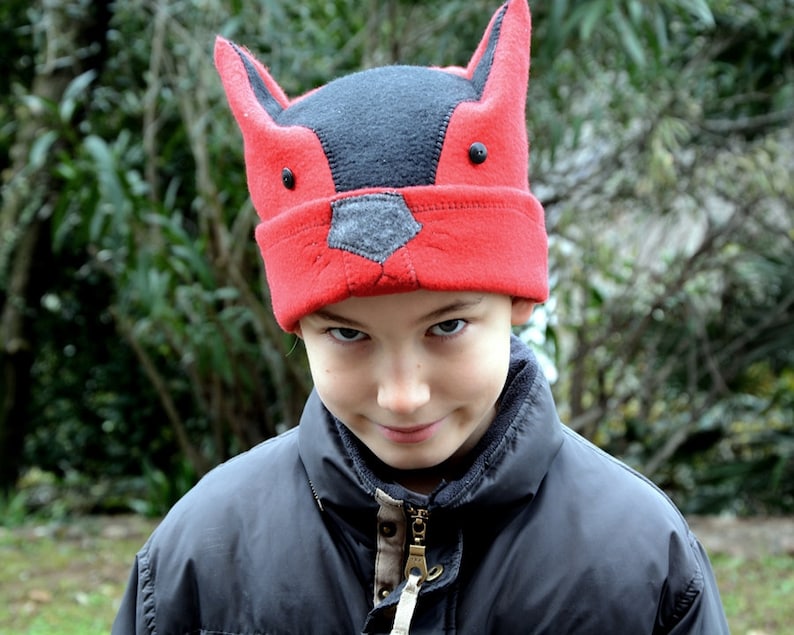 May include: A child wearing a red and black fox hat with a mischievous expression. The hat has large ears and a black nose.