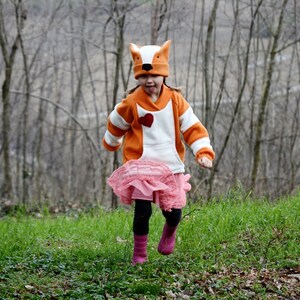 May include: A young person wearing a pink skirt and an orange and white fox hoodie with a red heart on the front. The person is wearing a fox hat and is running through a forest.