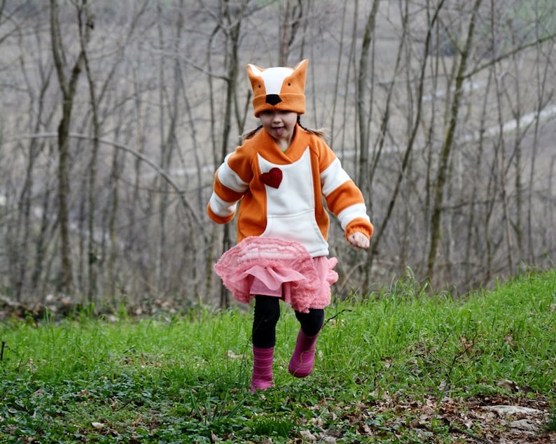 May include: A young person wearing a pink skirt and an orange and white fox hoodie with a red heart on the front. The person is wearing a fox hat and is running through a forest.