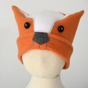 May include: Orange and white fleece hat with fox ears and black button eyes. The hat has a gray felt nose and stitched whiskers.