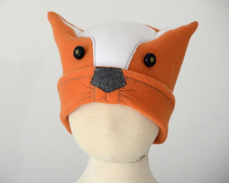 May include: Orange and white fleece hat with fox ears and black button eyes. The hat has a gray felt nose and stitched whiskers.