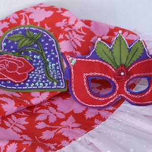 May include: A red felt mask with green leaves and a red rose detail. The mask is decorated with pink stitching and white beads. The mask is sitting on a pink and white fabric with a floral pattern.