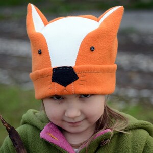 May include: A child wearing a bright orange and white fox hat with black felt eyes and nose. The hat has a white fur trim around the face opening. The child is wearing a green jacket with a pink lining.
