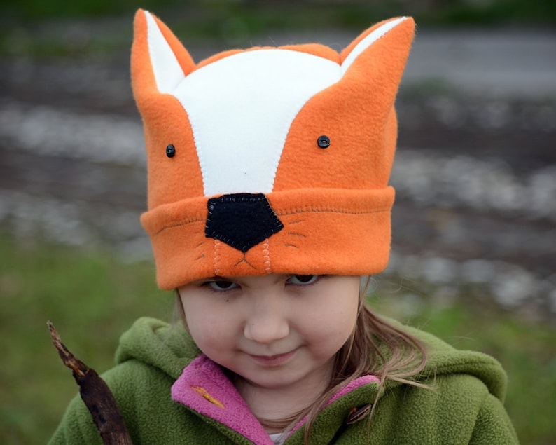 May include: A child wearing a bright orange and white fox hat with black felt eyes and nose. The hat has a white fur trim around the face opening. The child is wearing a green jacket with a pink lining.