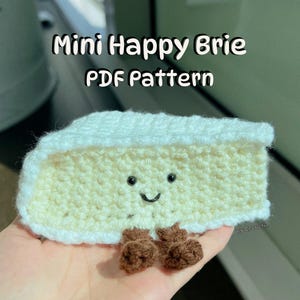 Puede incluir: Un patrón de amigurumi de crochet para un queso brie feliz. El patrón está disponible para descargar en PDF. El brie de crochet es blanco con un centro amarillo y tiene una cara sonriente. Tiene dos pies de crochet marrones.