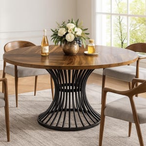 May include: A circular wooden dining table with a dark brown finish, supported by a black metal base. The table is set with a floral arrangement, bottles, and surrounded by wooden chairs with grey upholstered seats.