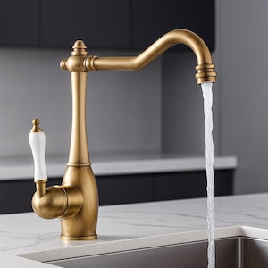 May include: A brass-finished kitchen tap with a curved spout and a white porcelain handle. Water flows from the spout into a stainless steel sink. The tap is mounted on a white countertop. The design is reminiscent of a vintage style.