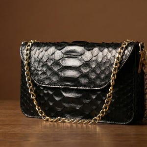May include: A black, textured leather handbag with a gold chain strap. The bag has a flap closure and a snakeskin pattern. The background is a warm brown color, and the bag is sitting on a wooden surface.