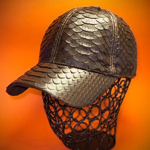 Könnte beinhalten: Eine braune Leder-Baseballkappe mit schuppiger Textur. Die Kappe befindet sich auf einem schwarzen Metallständer.