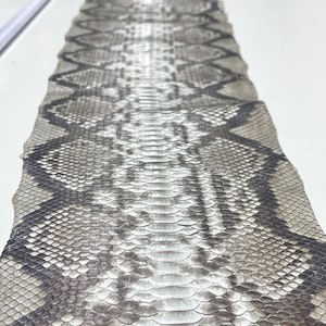 以下が含まれることがあります： グレーと黒の色合いのひし形模様のヘビ革のクローズアップ。鱗が見え、質感のある表面を作り出しています。この革は、工芸品や装飾目的で使用される可能性があります。