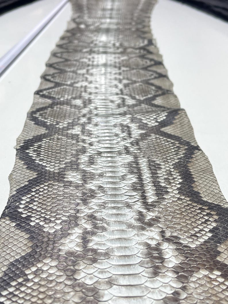 以下が含まれることがあります： グレーと黒の色合いのひし形模様のヘビ革のクローズアップ。鱗が見え、質感のある表面を作り出しています。この革は、工芸品や装飾目的で使用される可能性があります。