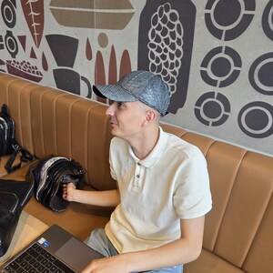 May include: A light gray baseball cap with a snakeskin pattern. The person is wearing a white polo shirt and blue jeans. A laptop and a black bag are on the table. The background features a brown and beige patterned wall.