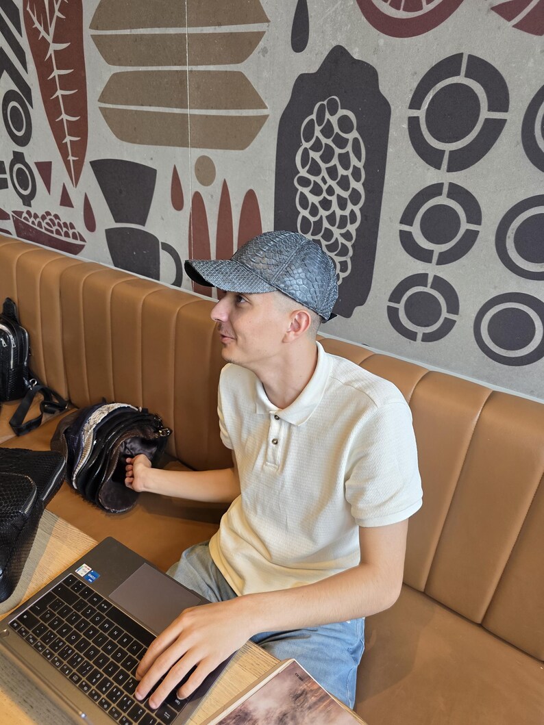 May include: A light gray baseball cap with a snakeskin pattern. The person is wearing a white polo shirt and blue jeans. A laptop and a black bag are on the table. The background features a brown and beige patterned wall.