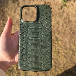 May include: Green textured phone case with a reptile skin pattern. The case has a rectangular camera cutout and a dark green border. The case is held against a background of dry grass and a brick wall.