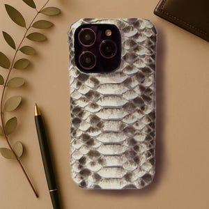 Könnte beinhalten: Eine weiße und graue Schlangenleder-Handyhülle mit einem schwarzen Kameraobjektiv-Ausschnitt.