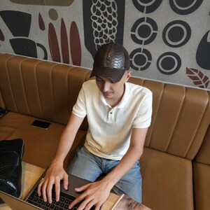 Op de afbeelding: Een bruine, getextureerde baseballcap met een reptielen huidpatroon. De pet wordt gedragen door een persoon die een witte poloshirt en een blauwe spijkerbroek draagt. De persoon zit aan een tafel met een laptop.