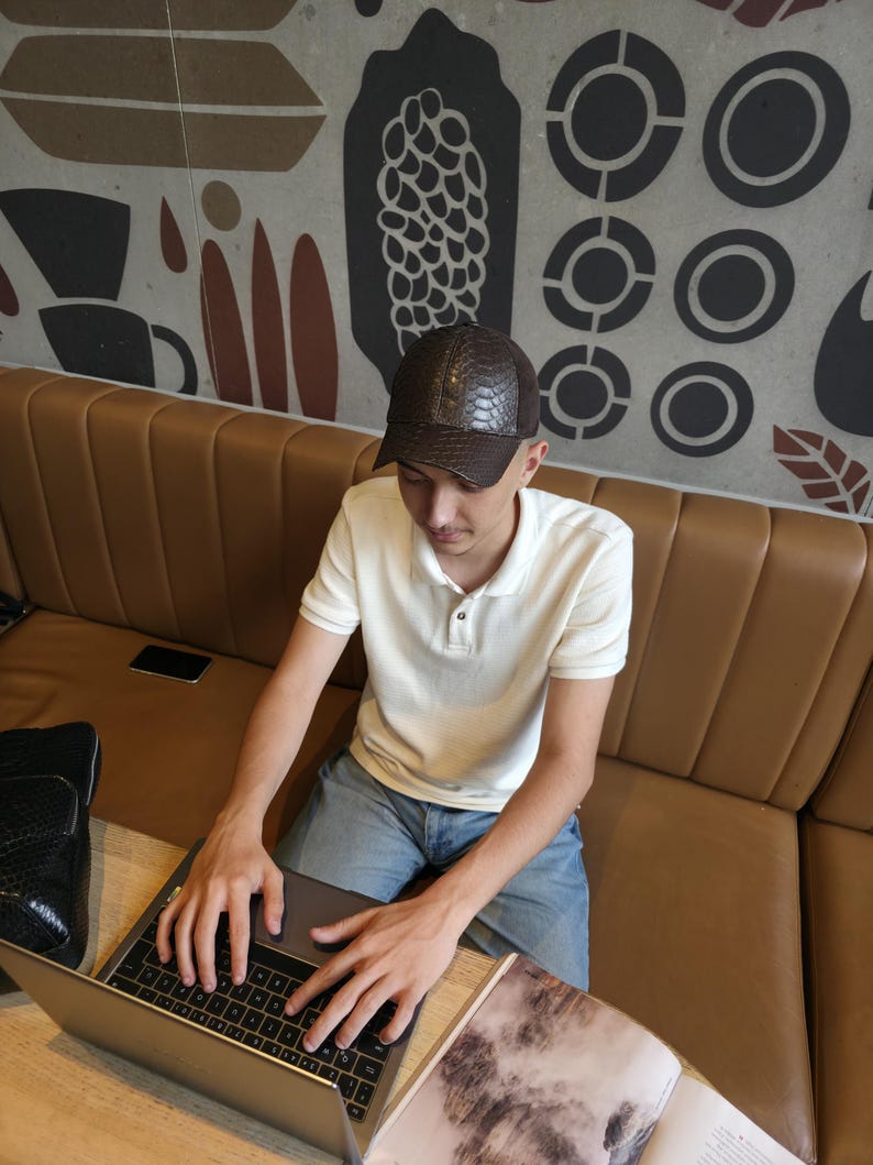 Op de afbeelding: Een bruine, getextureerde baseballcap met een reptielen huidpatroon. De pet wordt gedragen door een persoon die een witte poloshirt en een blauwe spijkerbroek draagt. De persoon zit aan een tafel met een laptop.