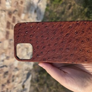 Pode incluir: Uma capa de telefone de couro castanho com um padr&atilde;o texturizado de avestruz. A capa tem formato retangular com cantos arredondados e um recorte para a c&acirc;mera. A capa &eacute; segurada em uma m&atilde;o.
