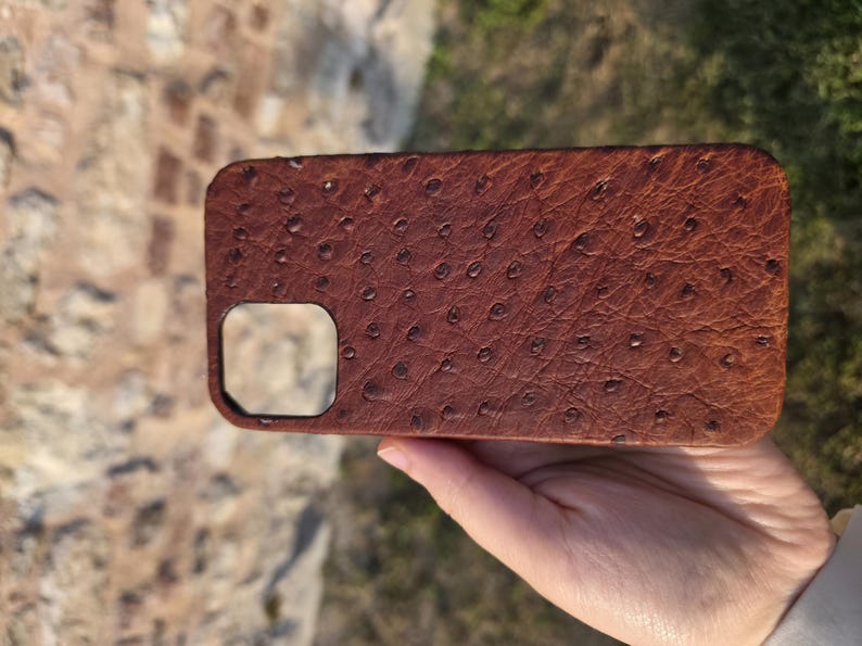 Pode incluir: Uma capa de telefone de couro castanho com um padr&atilde;o texturizado de avestruz. A capa tem formato retangular com cantos arredondados e um recorte para a c&acirc;mera. A capa &eacute; segurada em uma m&atilde;o.