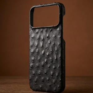 Op de afbeelding: Een zwarte telefoonhoes met een getextureerd oppervlak. De hoes heeft een rechthoekige opening voor de camera en een verhoogd, hobbelig patroon. De hoes is gemaakt van een donker materiaal en ligt op een houten oppervlak.