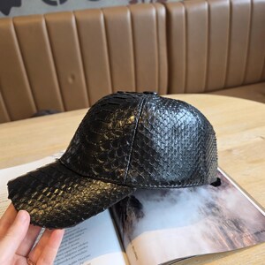 K&ouml;nnte beinhalten: Schwarze Baseballkappe mit einem strukturierten, schlangenlederartigen Muster. Die Kappe hat einen gebogenen Schirm und liegt auf einem offenen Magazin. Die Kappe ist dunkel und scheint aus Leder zu sein.