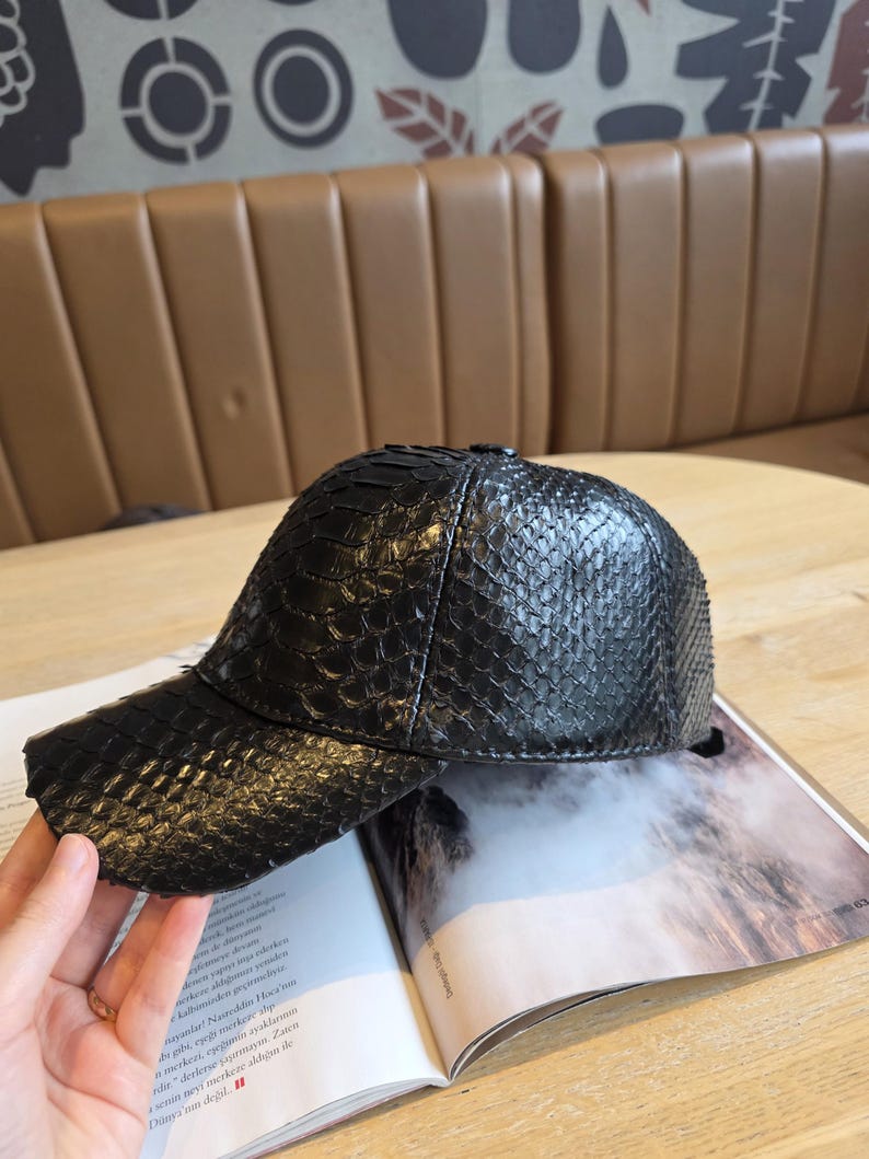 K&ouml;nnte beinhalten: Schwarze Baseballkappe mit einem strukturierten, schlangenlederartigen Muster. Die Kappe hat einen gebogenen Schirm und liegt auf einem offenen Magazin. Die Kappe ist dunkel und scheint aus Leder zu sein.