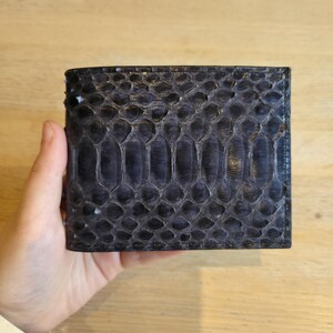 Puede incluir: Una cartera azul oscuro con un patr&oacute;n texturizado en forma de escamas. La cartera es rectangular y parece estar hecha de cuero. La cartera est&aacute; sujeta en la mano de una persona.