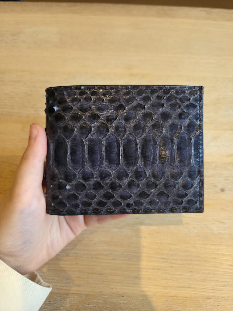 Puede incluir: Una cartera azul oscuro con un patr&oacute;n texturizado en forma de escamas. La cartera es rectangular y parece estar hecha de cuero. La cartera est&aacute; sujeta en la mano de una persona.