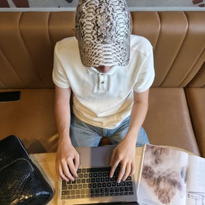 May include: A baseball cap with a snakeskin pattern in shades of gray, white, and black. The cap is worn by a person sitting at a table with a laptop and an open magazine. A black bag is also on the table.