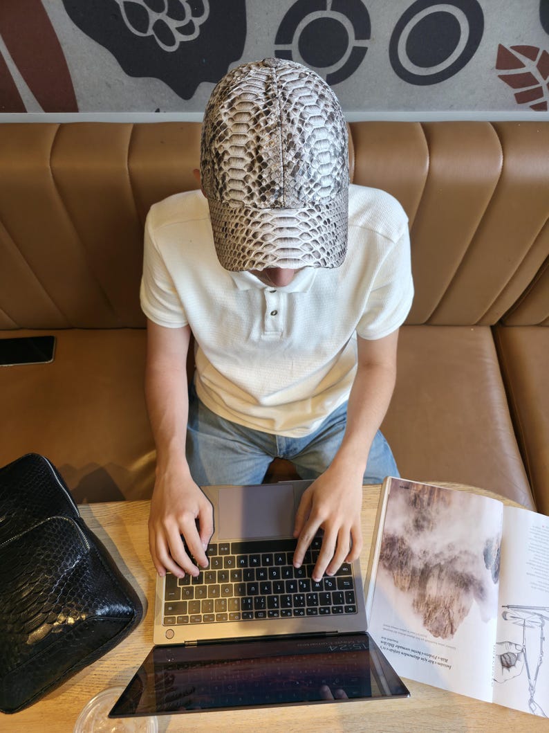 May include: A baseball cap with a snakeskin pattern in shades of gray, white, and black. The cap is worn by a person sitting at a table with a laptop and an open magazine. A black bag is also on the table.