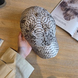 May include: A baseball cap with a snakeskin pattern in shades of gray and white. The cap has a curved brim and a structured crown. The hat is made of a textured material and is designed for casual wear.