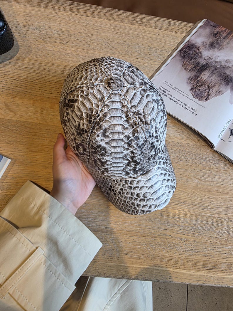 May include: A baseball cap with a snakeskin pattern in shades of gray and white. The cap has a curved brim and a structured crown. The hat is made of a textured material and is designed for casual wear.