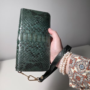 May include: A dark teal wallet with a snakeskin texture. The wallet has a wrist strap and a gold-colored clasp. The wallet is rectangular and appears to be made of leather. The wrist strap is black.