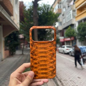 May include: Orange phone case with a snakeskin texture. The case has a rectangular shape with a cutout for the camera. The case is held in a person's hand, with a blurred background of a street and buildings.