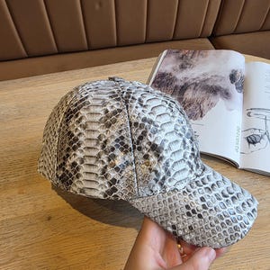 May include: A baseball cap with a snakeskin pattern in shades of gray and white. The cap is made of a textured material and has a curved brim. The cap is sitting on a wooden surface next to an open book.