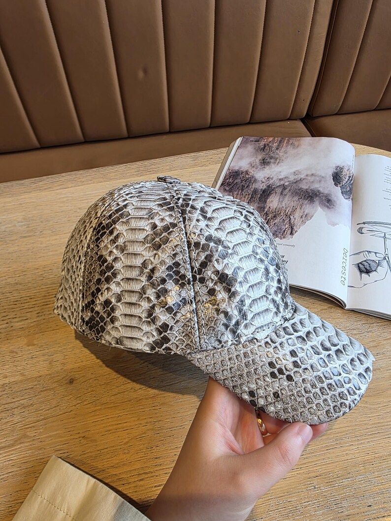May include: A baseball cap with a snakeskin pattern in shades of gray and white. The cap is made of a textured material and has a curved brim. The cap is sitting on a wooden surface next to an open book.