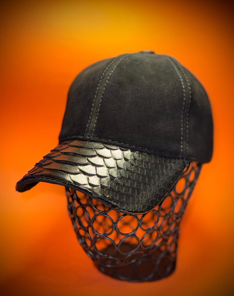 May include: A black baseball cap with a snakeskin-textured brim. The cap is displayed on a wire head-shaped form against an orange background. The cap's crown appears to be made of a soft, dark material.