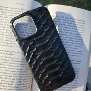 Könnte beinhalten: Schwarze, strukturierte Handyhülle mit schuppenartigem Muster, die auf einem offenen Buch liegt. Die Hülle hat Aussparungen für Kamera und Knöpfe. Die Seiten des Buches sind sichtbar, mit Text in einer Fremdsprache.