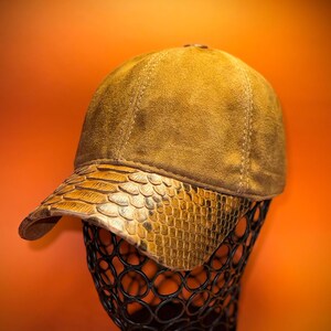 May include: A tan baseball cap with a snakeskin-textured brim. The cap is displayed on a black wire form against an orange background. The cap's crown is a solid tan color, and the brim has a scale-like pattern.