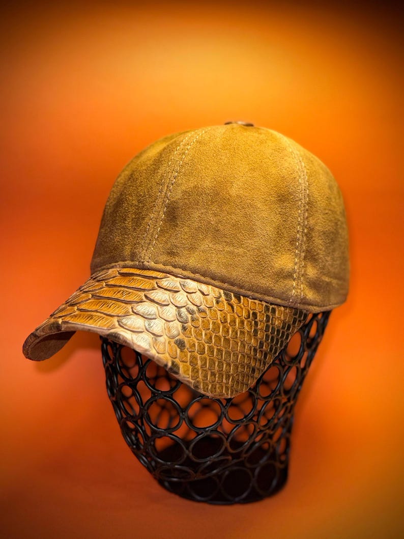 May include: A tan baseball cap with a snakeskin-textured brim. The cap is displayed on a black wire form against an orange background. The cap's crown is a solid tan color, and the brim has a scale-like pattern.