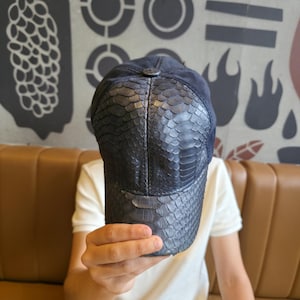Op de afbeelding: Een donkerblauwe baseballcap met een slangenpatroon op de klep en een suède kroon. De pet wordt voor een persoon omhoog gehouden. De achtergrond toont een bruine leren bank en een muur met abstracte ontwerpen.