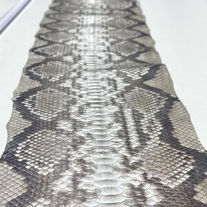 以下が含まれることがあります： グレーと黒の色合いのひし形模様のヘビ革のクローズアップ。鱗が見え、質感が粗いです。ヘビ革は、工芸品やファッションに使用される可能性があります。