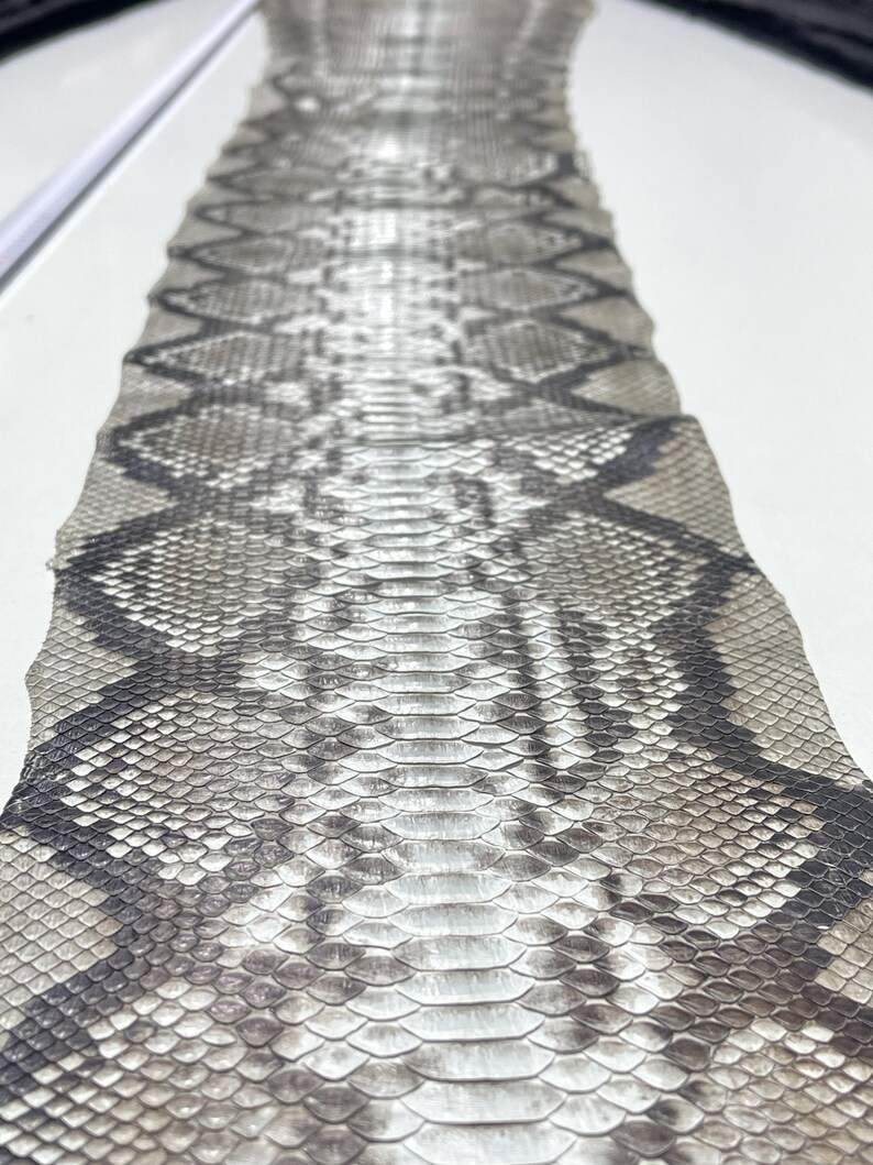 以下が含まれることがあります： グレーと黒の色合いのひし形模様のヘビ革のクローズアップ。鱗が見え、質感が粗いです。ヘビ革は、工芸品やファッションに使用される可能性があります。